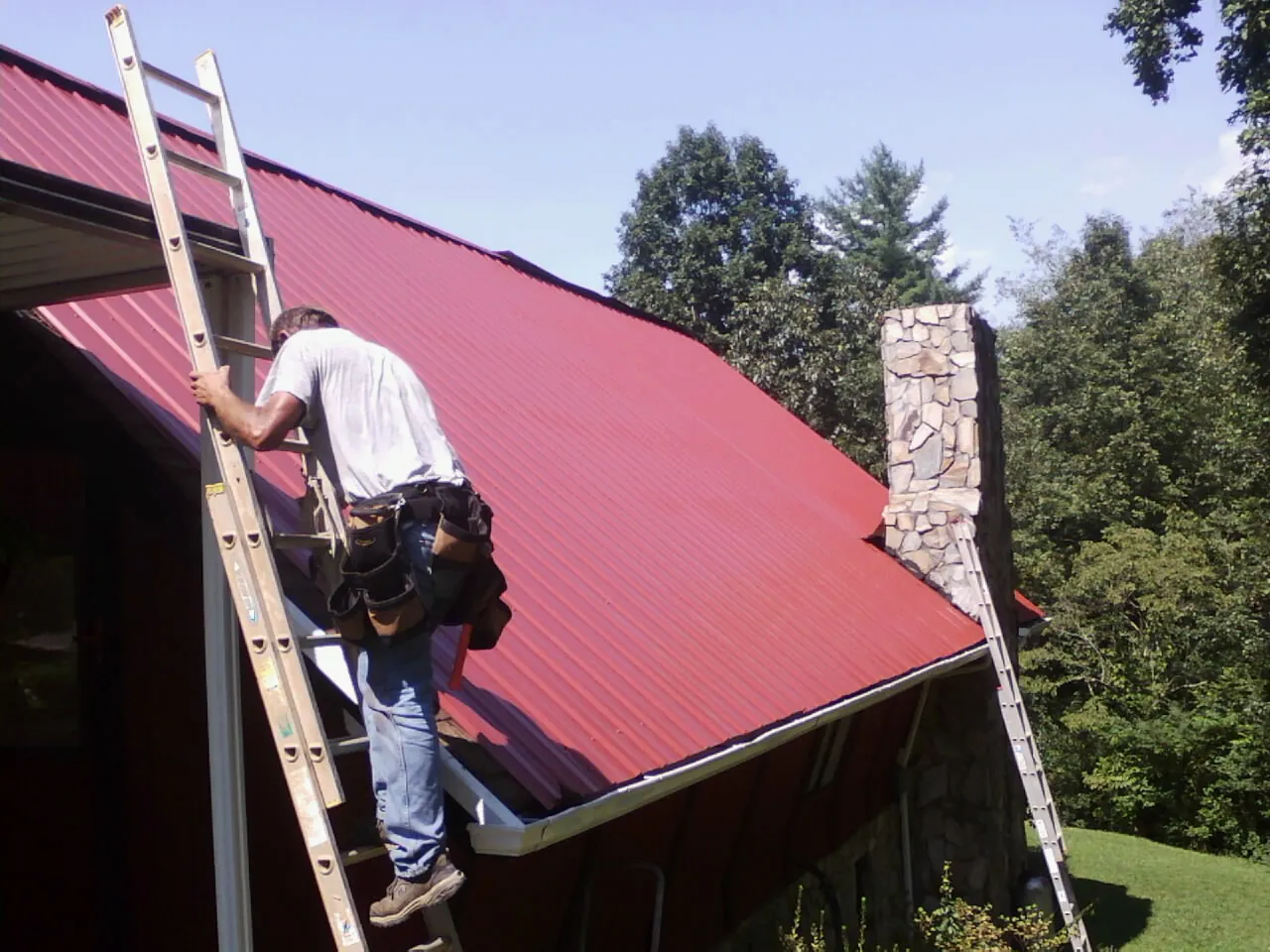 Roofer on a ladder beside a red metal roof and stone chimney — Soffit & Fascia Repair by Roofing Brothers in Milanville